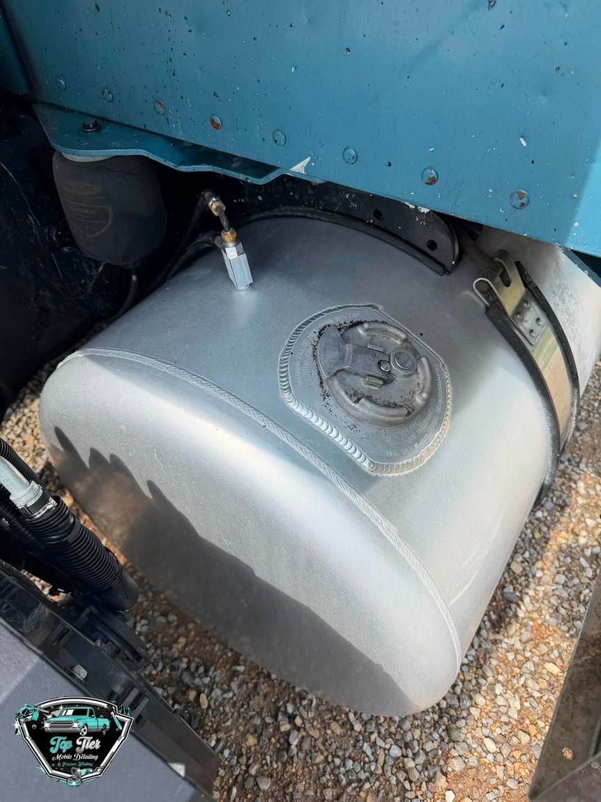 A wider view of a dull, dirty aluminum fuel tank under a truck cab
